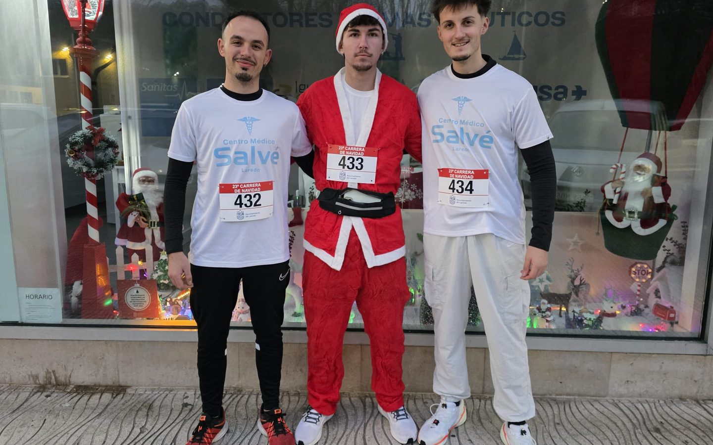 Tres amigos con dorsales delante de un escaparate navideño, uno vestido de Papá Noel